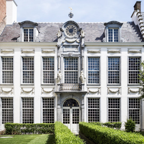 Elegantes historisches Gebäude mit symmetrischer Fassade, hohen, bogenförmigen Fenstern, verziertem Balkon und üppigen grünen Hecken im Vordergrund.
