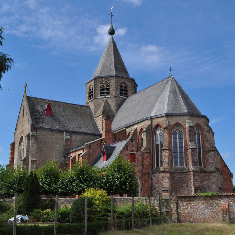 Eine historische Backsteinkirche mit hohem zentralen Turm und spitzem Dach, umgeben von üppigem Grün, unter einem klaren blauen Himmel, die eine ruhige Atmosphäre vermittelt.