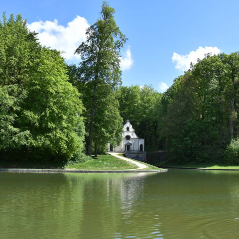 Ruhige Parkszene mit einer kleinen weißen Kapelle, umgeben von üppigen grünen Bäumen, die sich in einem stillen Teich unter strahlend blauem Himmel spiegeln. Enten schwimmen im Vordergrund.