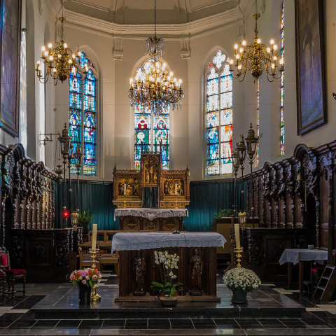 Innenraum einer prächtigen Kirche mit kunstvollen Holzschnitzereien, einem zentralen Altar geschmückt mit Blumen und farbenfrohen Buntglasfenstern. Elegante Kronleuchter schaffen eine ruhige und ehrfürchtige Atmosphäre.