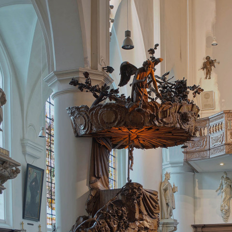 Eine kunstvoll geschnitzte Holzkanzel im Kircheninneren, geschmückt mit Figuren und filigranen Details. Sonnenlicht fällt durch Buntglas und schafft eine ruhige Atmosphäre.