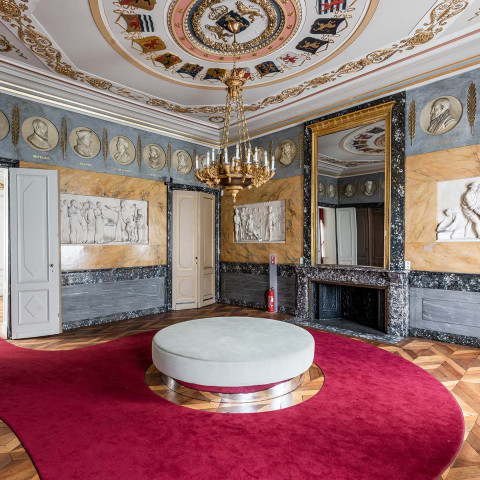 Prächtiger Raum mit reich verziertem Deckenfries mit runden Porträts, großem Kronleuchter, rotem Teppich, zentralem runden Ottomane und Wänden mit klassischen Reliefs.