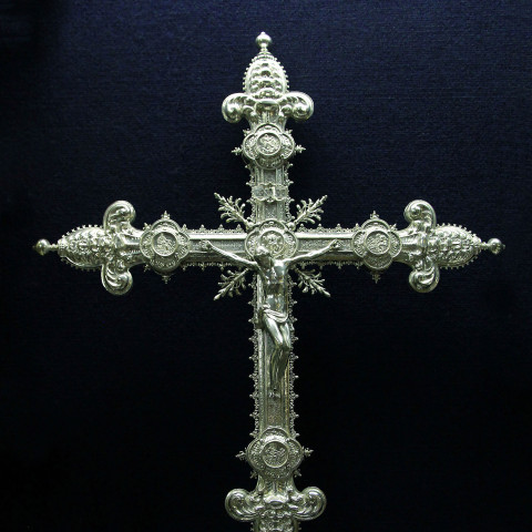 Aufwändig verzieltes silbernes Kreuz vor dunklem Hintergrund, mit filigranen Mustern und einer zentralen Christusfigur. Der Ton ist feierlich und ehrfürchtig.