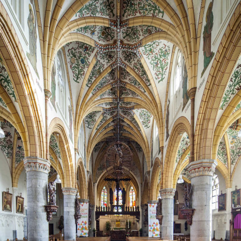 Weitläufiges Kircheninnere mit hohen, verzierten Bögen, detaillierter Gewölbedecke und Reihen hölzerner Kirchenbänke. Buntglasfenster verleihen lebendige Farben.