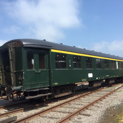 Vintage grüner Eisenbahnwagen mit gelbem Streifen auf den Gleisen unter blauem Himmel. Üppiges Grün umgibt die Szene und vermittelt ein Gefühl von Nostalgie und Ruhe.