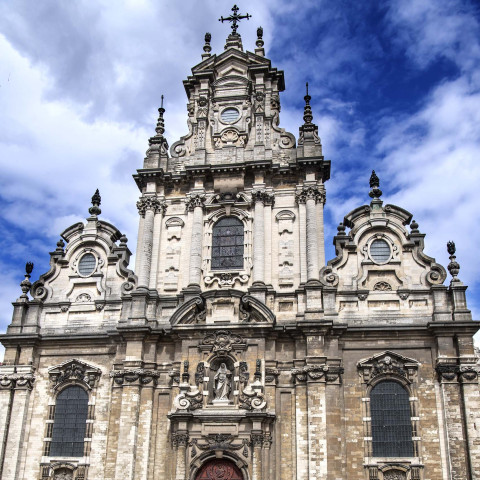 Baroque-style church façade with ornate stone carvings, arched windows, and a cross at the top against a vibrant blue sky with white clouds.