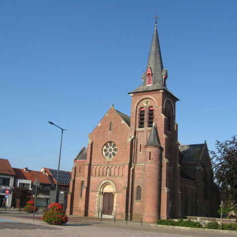 Eine historische Kirche aus rotem Backstein mit hohem Turm und Rundfenster steht unter einem klaren blauen Himmel, umgeben von kleinen Gebäuden und Bäumen.