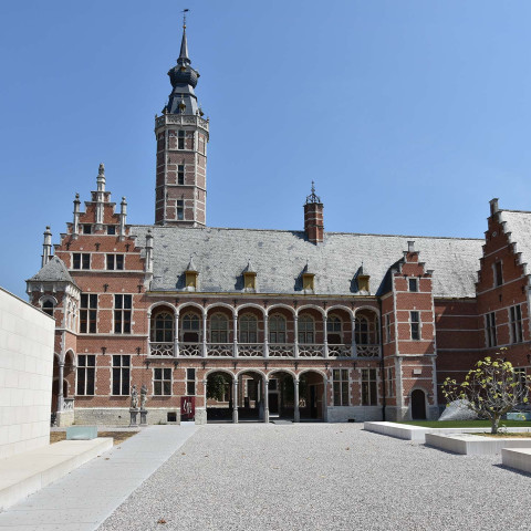 Historic brick building with ornate gables and a tall clock tower under a clear blue sky. The foreground features a spacious cobblestone courtyard.