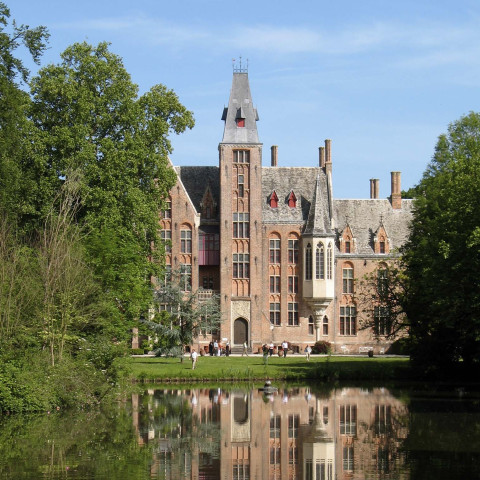 Ein historisches Backsteinschloss mit hohen Türmen und verzierten Fenstern steht an einem ruhigen See. Bäume umgeben die Szene und spiegeln sich auf der Wasseroberfläche bei klarem blauen Himmel.