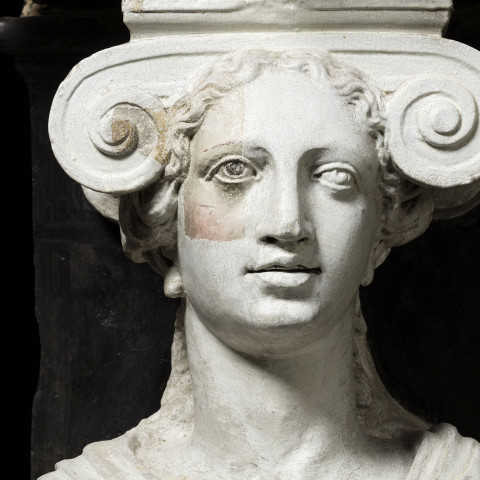 Bust of a woman with an ornate, scroll-like headdress, gazing outward. The sculpture is white marble with soft facial features, conveying a serene expression.