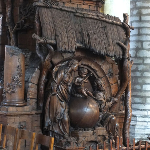 Ornate wooden relief depicting Mary, baby Jesus, and Joseph under a textured canopy. The scene, rich with intricate details, conveys a serene, sacred mood.