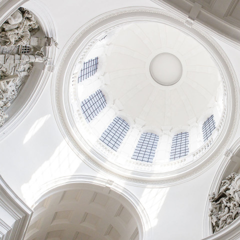The image shows a majestic white domed ceiling of a church interior. Intricate sculptures adorn the curved walls, and arched windows let in soft light.