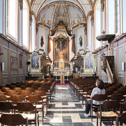 Eine Frau sitzt allein in einer historischen Kirche mit kunstvollem Gewölbe, hölzernen Kirchenbänken und einem aufwendig gestalteten Altar, was eine ruhige, nachdenkliche Atmosphäre vermittelt.