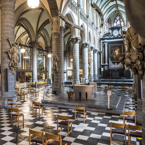 Grand cathedral interior with checkered floor, ornate columns, and arched ceilings. Wooden chairs spaced apart create a solemn and serene atmosphere.