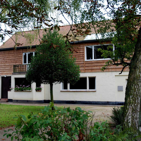 Ein zweistöckiges Haus mit Holzverkleidung und weißen unteren Wänden, eingebettet in einen üppigen Garten. Bäume rahmen die Szene ein und schaffen eine stille, einladende Atmosphäre.