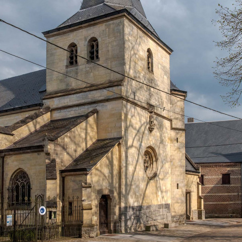 Steinkirche mit hohem, spitzem Turm unter einem bewölkten Himmel. Schatten von Bäumen fallen auf die hellfarbene Fassade und schaffen eine ruhige, kontemplative Atmosphäre.