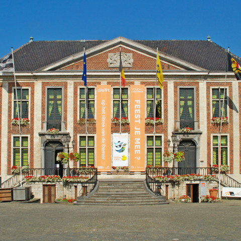 Historic building with a symmetrical facade, large windows, and decorative columns. Flags and banners adorn it, with steps leading to the entrance. Clear, sunny sky.