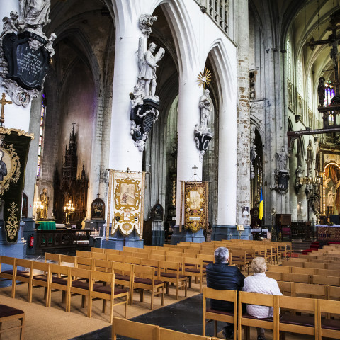 A grand, ornate cathedral interior with high arches, stained glass, intricate statues, and gilded altar. Two people sit on wooden pews, evoking a serene atmosphere.