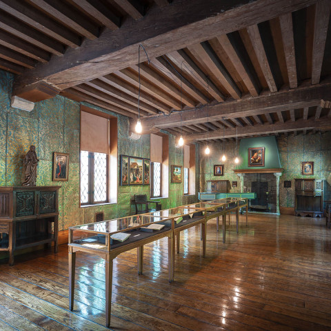 Dimly lit historic room with wooden beams, polished floors, and display cases. Medieval décor with portraits and statues creates a serene, scholarly ambiance.