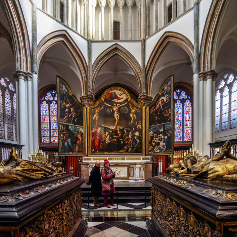 Innenraum einer Kathedrale mit zwei Personen, die einen Altar mit einem großen Kreuzigungsgemälde betrachten. Umgebende Buntglasfenster erzeugen farbiges Licht.