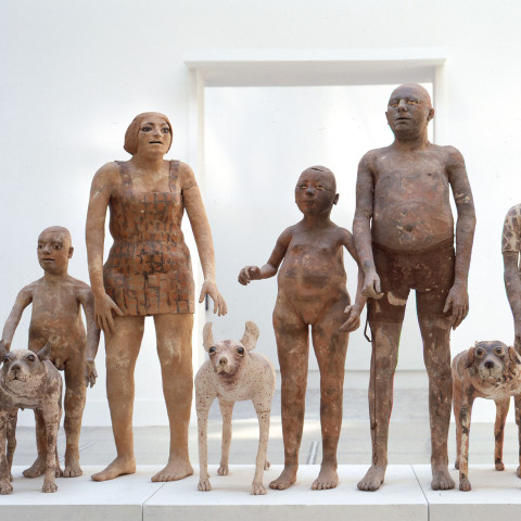 A row of clay statues depicting diverse human forms and dogs stands against a white background, conveying a sense of unity and artistic expression.