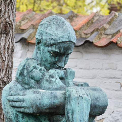 Bronzestatue einer Mutter, die ihr Kind zärtlich in den Armen hält. Vor einer rustikalen weißen Backsteinwand und Bäumen platziert, vermittelt sie Liebe und Schutz.