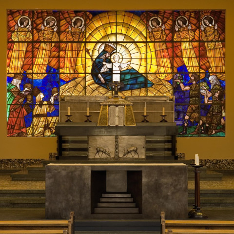 Buntglasfenster zeigen eine lebendige Weihnachtsgeschichte mit Maria und Jesus, umgeben von Engeln. Der Altar vorne verstärkt die ruhige, sakrale Atmosphäre.