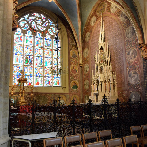 Interior of a church with ornate stained glass windows, decorative wall panels, and elaborate altar. Rows of wooden chairs create a serene atmosphere.