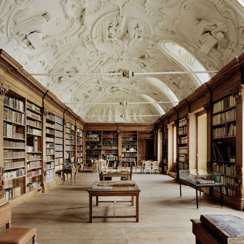 Spacious, ornate library with high, intricately decorated ceilings and wooden bookshelves lining the walls. Tables and chairs create a serene, scholarly atmosphere.