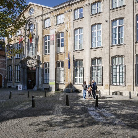 Historisches Steingebäude mit Bogenfenstern und dekorativem Wappen an der Fassade. Menschen gehen durch einen sonnendurchfluteten Kopfsteinpflasterhof mit Schatten.